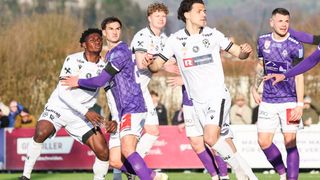 Fußballspieler in weißen und lila Trikots sind auf einem Feld. Der Spieler in Lila hat seinen Arm um den Spieler in Weiß. Ein anderer Spieler in Weiß beobachtet sie.