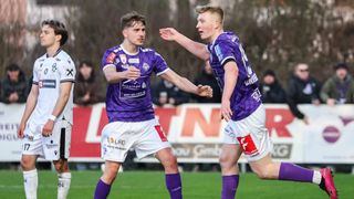 Zwei Fußballspieler in lila und weißen Trikots feiern ein Tor auf dem Feld, während ein dritter Spieler in Weiß hinter ihnen steht.