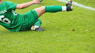 Ein Fußballspieler in Grün liegt auf dem Rasenfeld. Der Ball fliegt hinter ihm. Der Fuß des Spielers ist in der Luft gehoben.