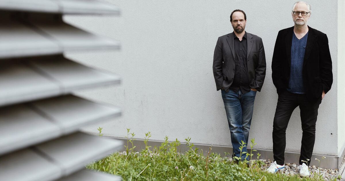 Ein Mann mit Bart steht mit den Händen in den Taschen vor einer weißen Wand, neben einem anderen Mann in Schwarz.