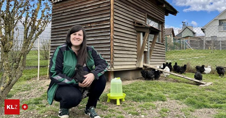 Eine Frau hält ein Küken in den Händen und steht neben einem Hühnerstall. Mehrere Hühner sind vor dem Stall.