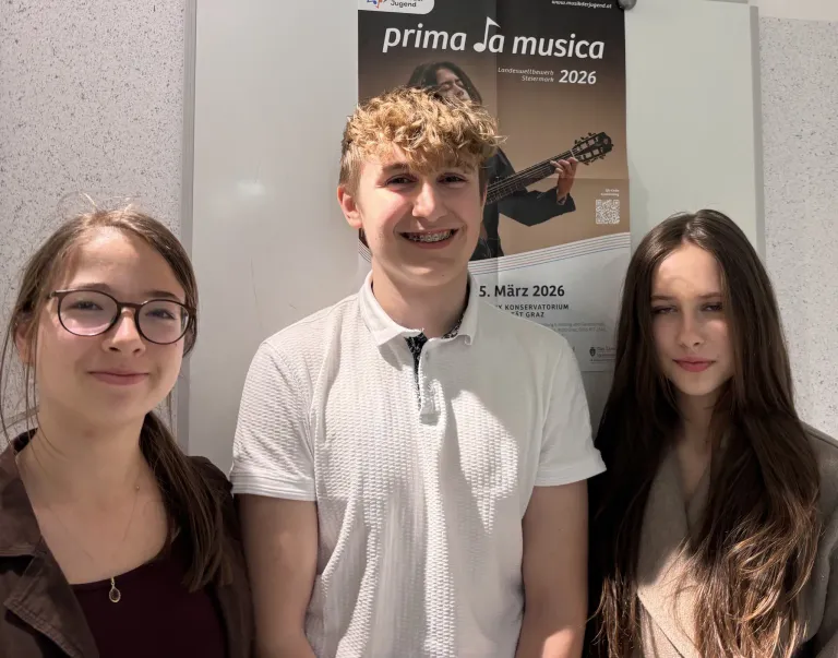Three people stand in front of a poster, a boy in the middle smiles with a guitar player on the poster above, two girls on either side.