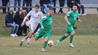Zwei Fußballspieler rennen auf einem Feld, einer in Grün und einer in Weiß, mit einem dritten Spieler und Zuschauern im Hintergrund.