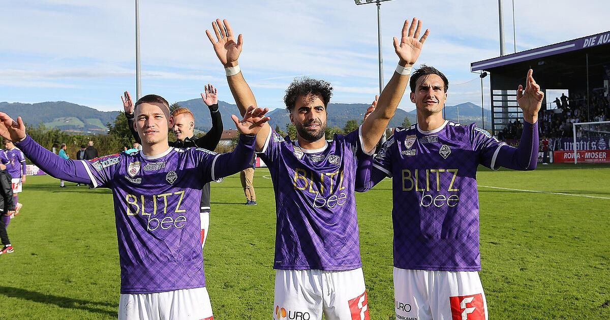 Drei Fußballspieler in lila Trikots mit 'BLITZ' und 'bee' auf der Vorderseite heben ihre Hände auf einem Rasenfeld.