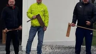 Drei Männer stehen im Freien, einer in einer gelben Jacke, der andere in einer schwarzen Jacke, beide halten Holzstäbe. Sie posieren für ein Foto vor einem weißen Gebäude.