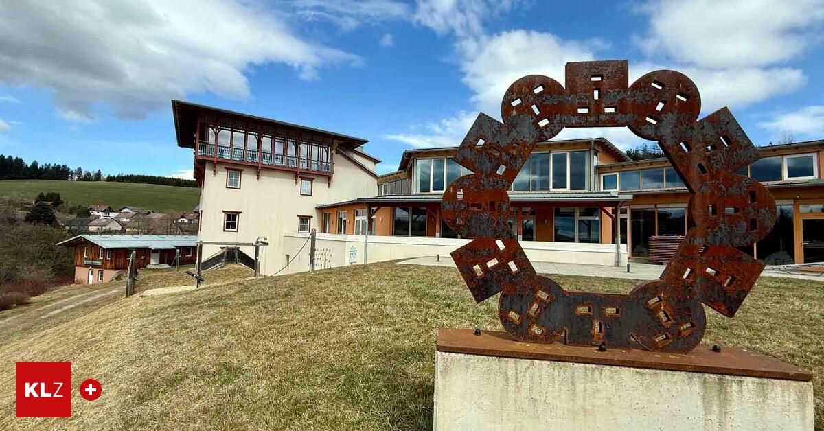 A modern building with a balcony and many windows stands on a hill with a grassy field in front. In the foreground, there is a rusty metal sculpture.