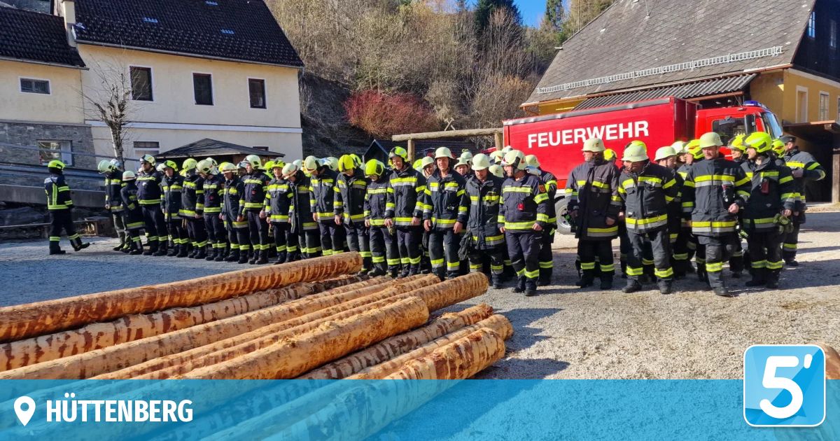 A line of firefighters in uniform, some standing and some sitting, with logs on the ground in front of them. A red fire truck is behind them.
