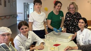 Mehrere Personen in einem Raum, eine trägt ein grünes Shirt mit Logo, eine andere eine Kochuniform, und andere lächeln. Ein Tisch mit einem Tablett, Gläsern und Teig. Eine gelbe Ballon im Hintergrund.