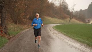 Ein älterer Mann joggt auf einer nassen, kurvenreichen Straße, umgeben von Bäumen und Gras. Er trägt ein blaues Shirt und schwarze Shorts.