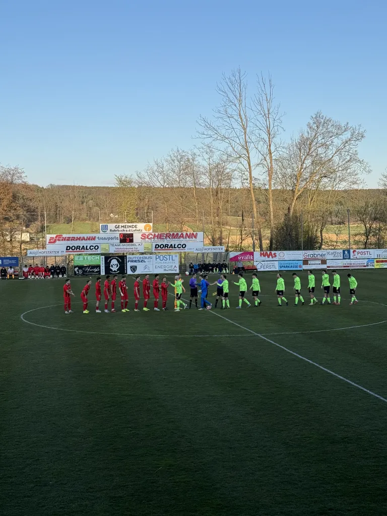 Soccer players in red and green uniforms line up on the field, possibly preparing for a match. Behind them, several advertisement banners are visible, and spectators are seated in the background.