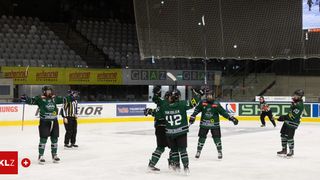 Eine Eishockeymannschaft feiert vor einem Tor, ein Spieler hält einen Schläger. Die Arena hat leere Tribünen, Banner und ein großes Netz.
