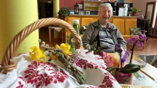 Eine ältere Frau sitzt in einem Restaurant mit einem Korb voller Blumen und einer Flasche vor sich. Der Raum hat einen Tisch, ein Regal mit Gegenständen und eine Wand mit Bildern.