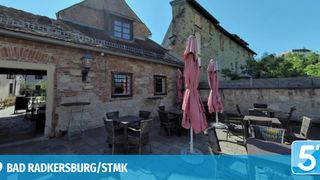 Ein Outdoor-Cafe mit Ziegelwänden, mit Tischen und Stühlen unter rosa Sonnenschirmen. Das Café hat einen Vintage-Charme mit einer Lampe an der Wand.