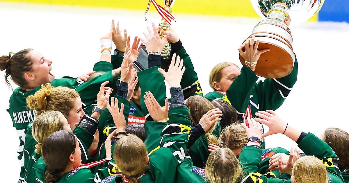 Eine Frauen-Eishockeymannschaft feiert mit einem Pokal in einer Eishalle, umgeben von jubelnden Teamkolleginnen.