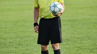 Ein Fußballschiedsrichter steht auf einem Feld, hält einen Fußball in einer Hand und eine gelbe Karte in der anderen. Er trägt ein neongelbes Trikot und schwarze Shorts.