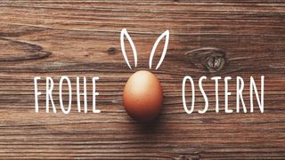 Ein braunes Osterei liegt auf einer Holzoberfläche, umgeben von Hasenohren und den Worten Rohe und Ostern in weiß.