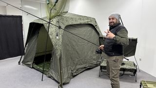 Ein Mann in einer grünen Mütze und Jacke steht neben einem getarnten Zelt und hält eine Angelrute mit einem Netz. Das Zelt ist aufgebaut und befindet sich in einem Raum.