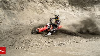 Ein Motocross-Fahrer fährt auf einer roten Maschine und wirft Staub auf einer Schotterpiste auf. Der Fahrer trägt einen Helm und einen Rennanzug.