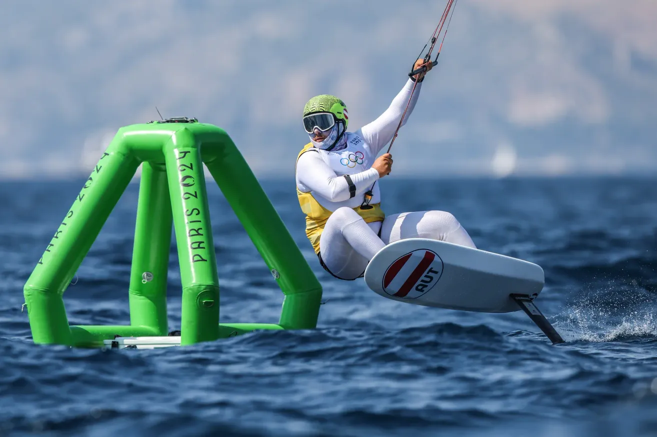 Ein Kiteboarder in weißer Ausrüstung mit dem Olympischen Logo nimmt an einer Ozeanveranstaltung teil. Grüne aufblasbare Objekte sind dahinter.