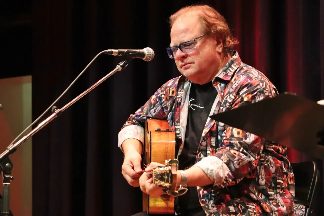 Ein Mann steht mit einer Gitarre auf der Bühne und singt in ein Mikrofon. Er trägt eine Brille und ein buntes Shirt.