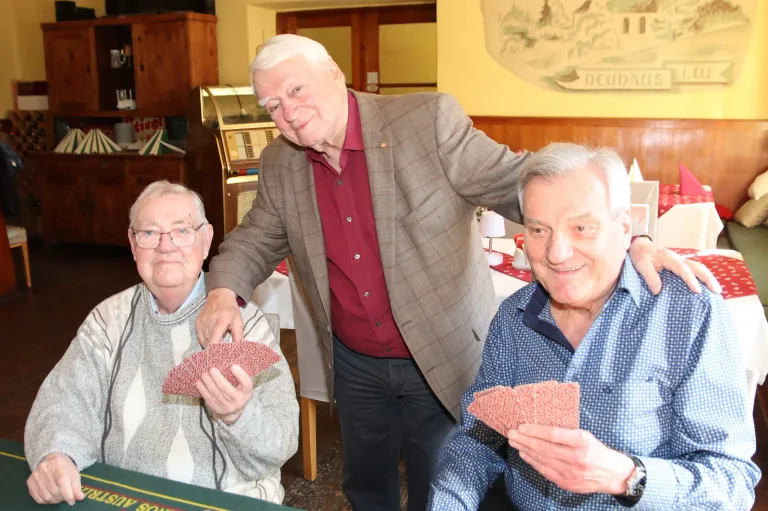 Three elderly men sit at a table, two of them holding cards, and the third man standing behind them. They all look cheerful. The room has a jukebox, wooden furniture, and a wall with a painting.