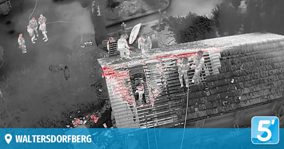 Ein Schwarz-Weiß-Bild zeigt ein beschädigtes Gebäude mit Feuerwehrleuten auf dem Dach, einer hält eine Leiter und der andere eine Seil. Das Gebäude ist von Wasser umgeben.