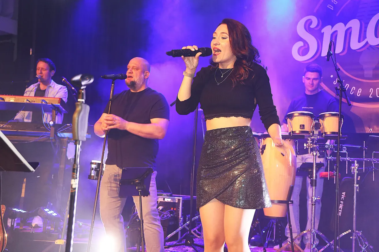 Eine Frau singt in ein Mikrofon auf der Bühne mit einem Mann und einer Band. Die Bühne ist mit lila Lichtern beleuchtet und es gibt Trommeln hinter ihnen.