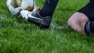 Ein leicht verschmutztes Fußballspielgerät ruht auf Gras. Ein Fuß des Spielers in einem Nike-Fußballschuh ist in der Nähe.