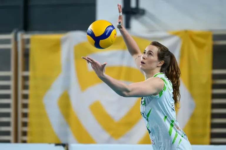 Eine Volleyballspielerin in einem grünen Trikot versucht, einen gelb-blauen Ball in der Luft zu treffen. Der Hintergrund zeigt ein gelbes Banner mit einem weißen 'C'.