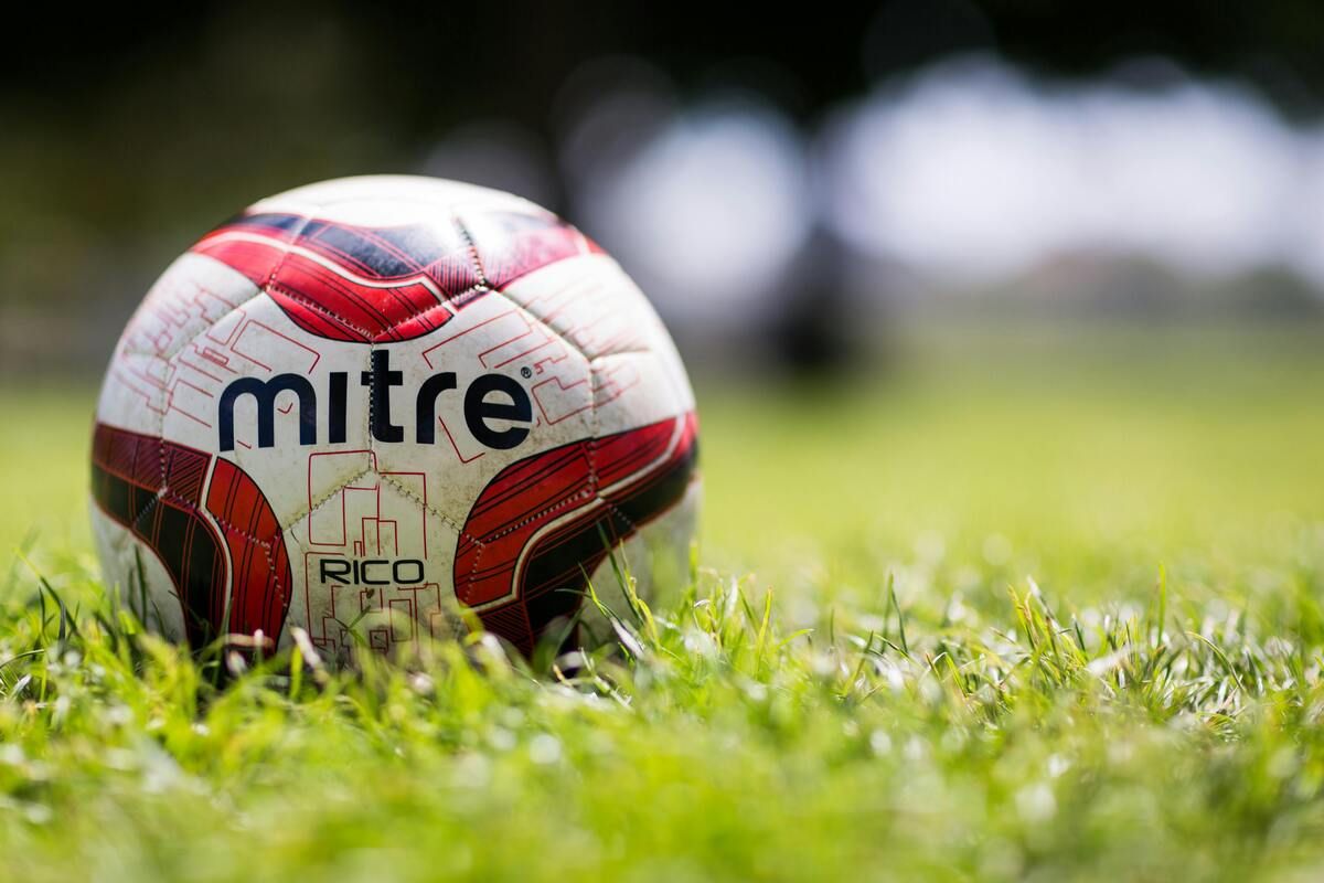A red and white soccer ball with the word 'Mitre' and 'RICO' printed on it is on the grass. The background is blurry.