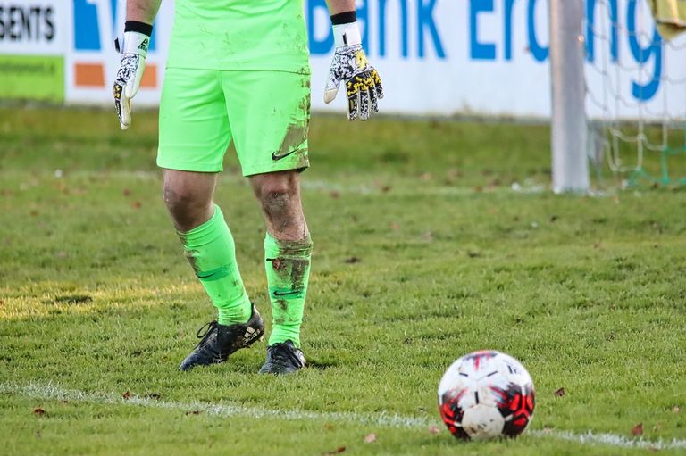 Ein Fußballtorwart steht auf einem Rasenfeld, trägt ein neongrünes Trikot und Handschuhe und hat Schlamm an den Schienbeinen. Ein Fußball mit roten und schwarzen Markierungen liegt in der Nähe.