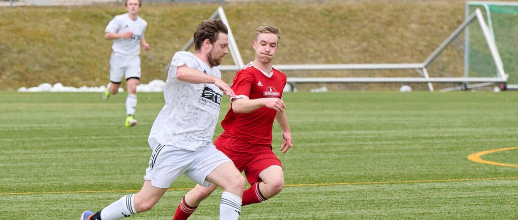 Zwei Fußballspieler in roten und weißen Trikots konkurrieren auf einem Feld. Ein Spieler ist dabei, den Ball zu schießen, während der andere verteidigt.