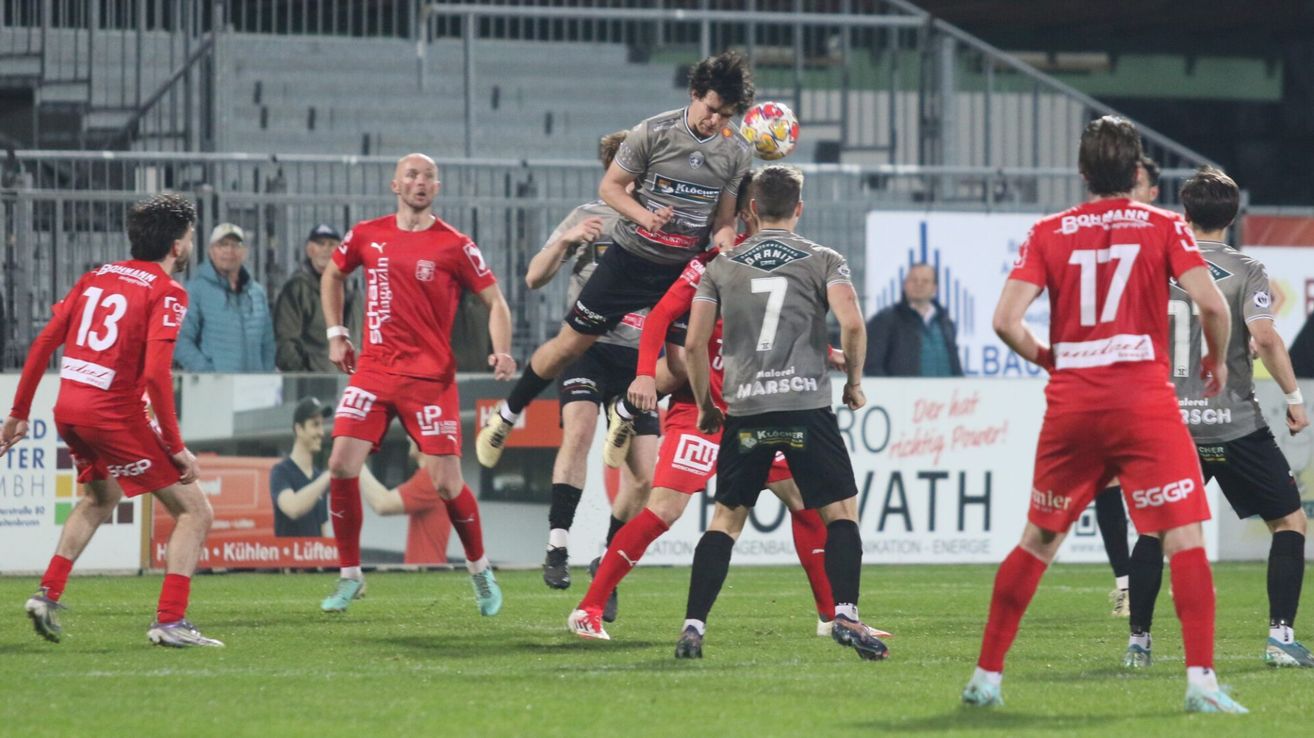 Fußballspieler in roten und grauen Trikots kämpfen um einen Ball in der Luft. Ein Spieler im roten Trikot läuft links, während andere springen und sich in der Luft strecken.