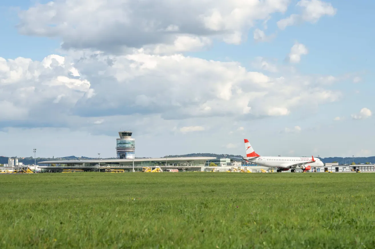 A red and white airplane is parked on the tarmac at an airport. A large control tower is visible behind it. The runway is empty with lush green grass. The sky is blue with scattered clouds.