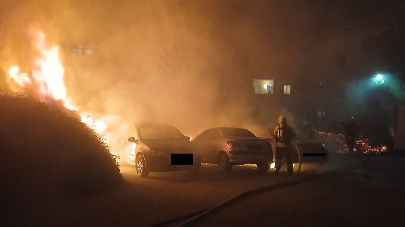 Ein Feuerwehrmann steht neben einem brennenden Auto und löscht die Flammen mit einem Schlauch. Rauch erfüllt die Umgebung, und ein zweites Auto ist in der Nähe geparkt.
