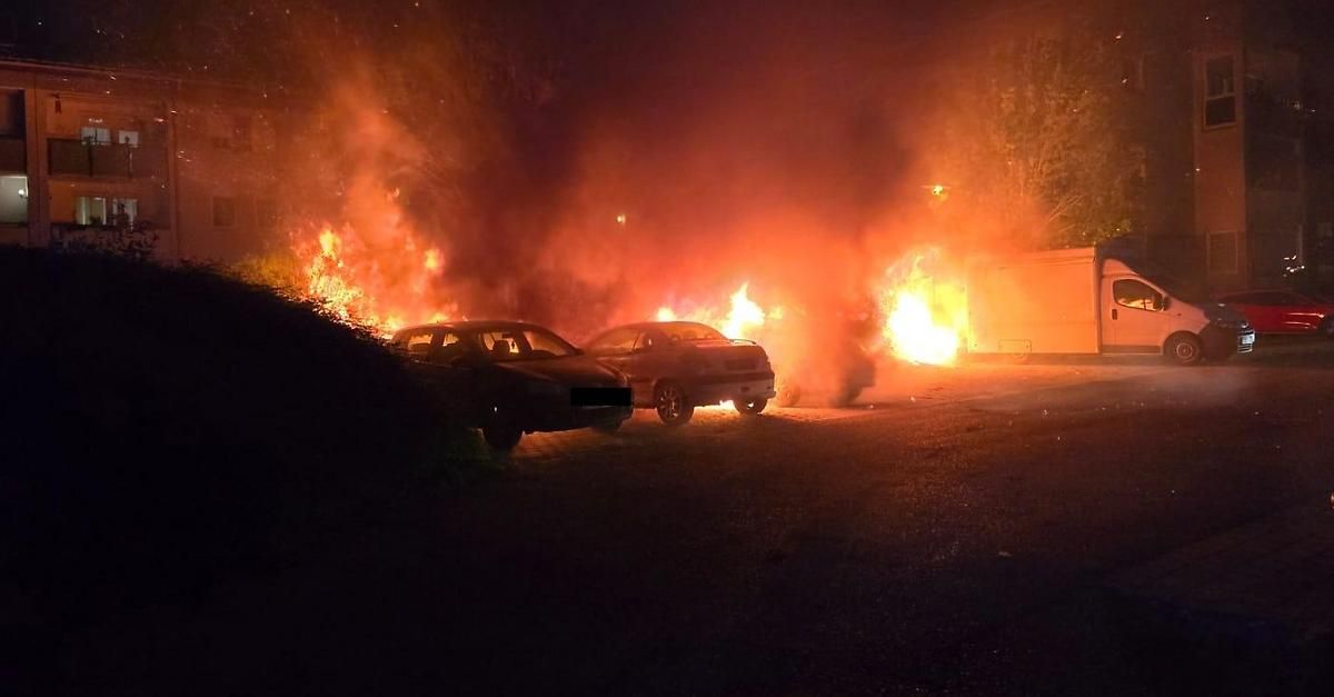 Zwei Autos auf einer dunklen Straße stehen in Flammen. Das Feuer breitet sich auf der Straße aus, und Rauch füllt die Luft.