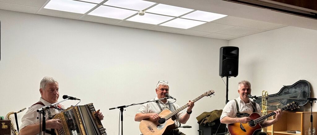 Drei Männer spielen Musik in einem Raum. Ein Mann auf der linken Seite spielt Akkordeon. In der Mitte spielt ein Mann Gitarre. Auf der rechten Seite spielt ein anderer Mann Gitarre und Trompete.