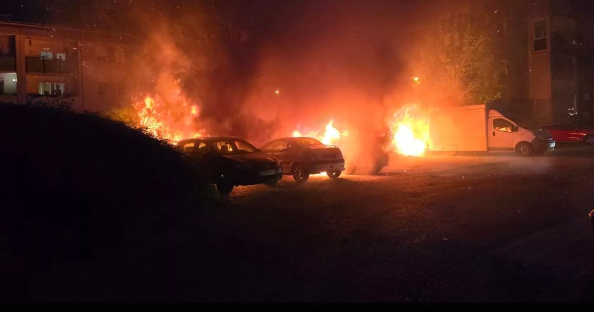Zwei Autos sind in der Nähe eines großen Feuers geparkt, das eine orangefarbene Glut auf die Nachtszene wirft.