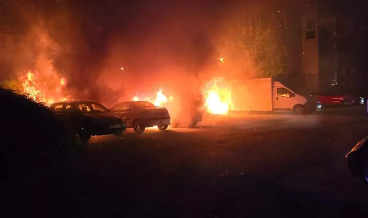 In der Nacht brennt ein Feuer einen weißen Lieferwagen und zwei draußen geparkte Autos. Die Flammen sind intensiv, und die Szene ist dunkel.