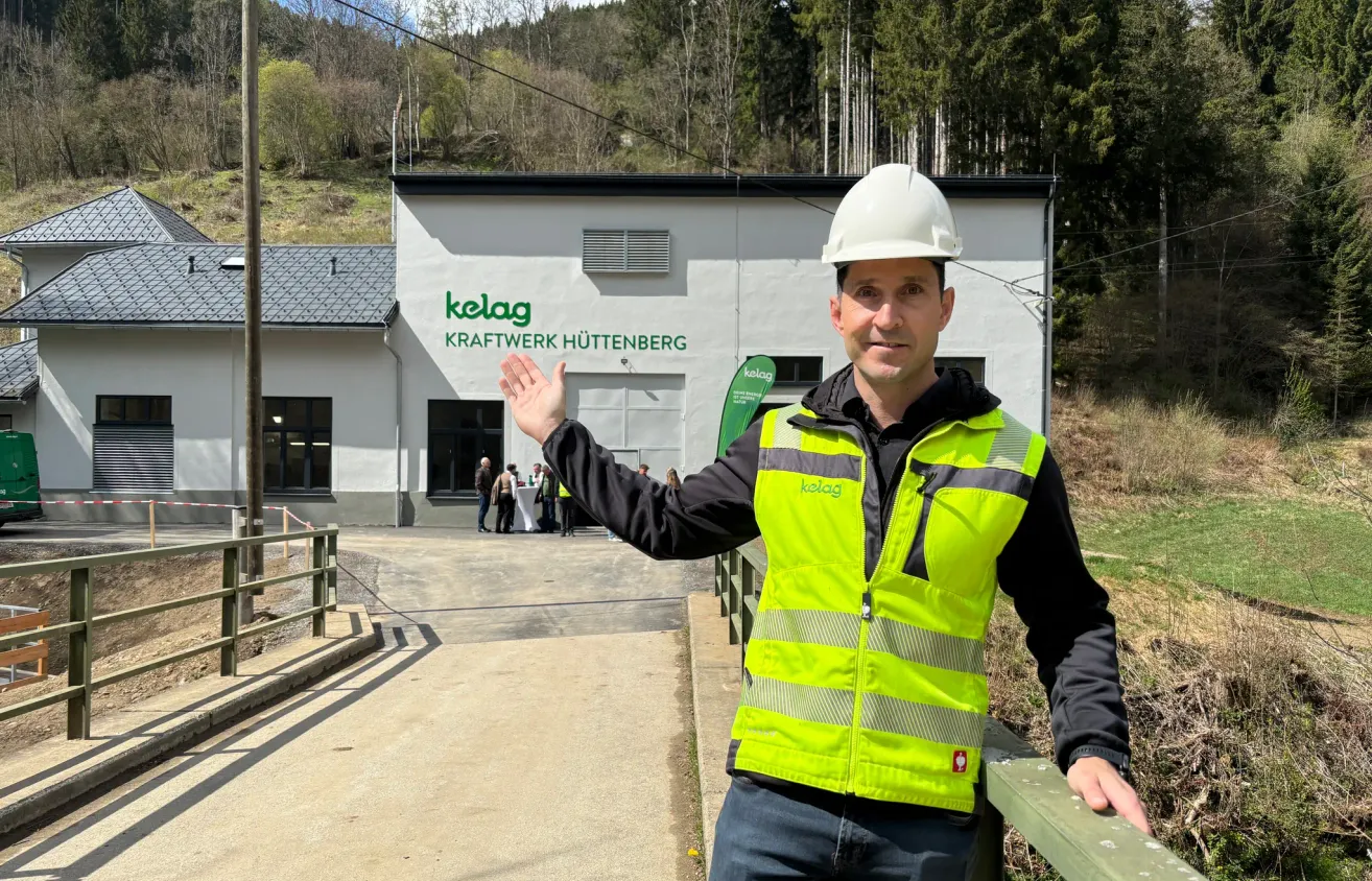 A man wearing a safety vest and hard hat is waving in front of the Kraftwerk Huttenberg building.