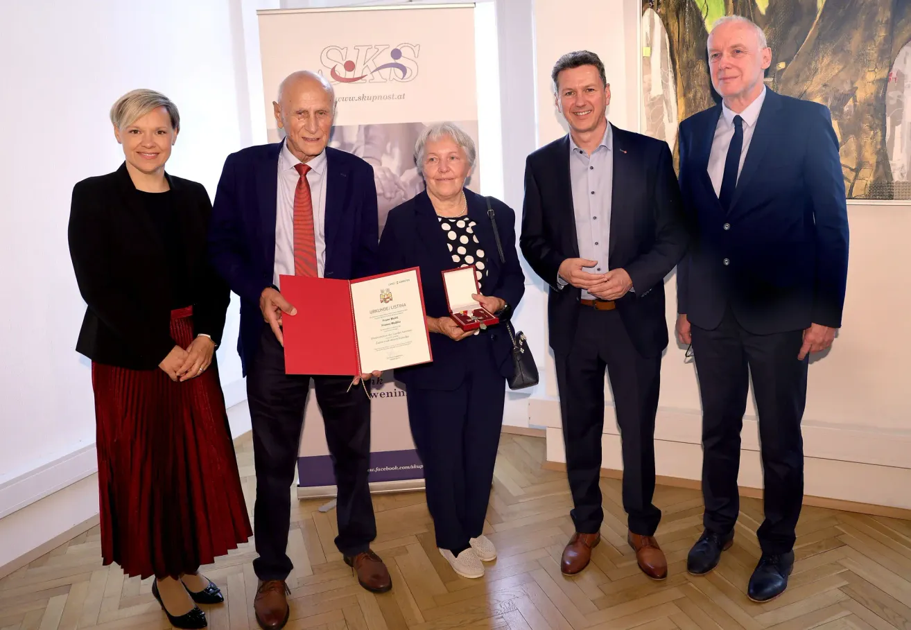 Five people stand together holding certificates. A woman in the center holds a red book and a box. The man next to her smiles. The man on the right wears a tie. Behind them is a banner and a painting.