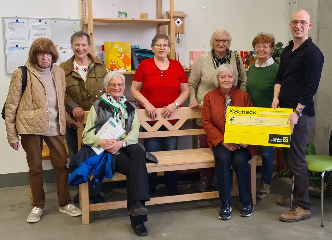 Seven women pose for a photo, holding a large check for 2,000 euros. They sit on a wooden bench and smile. Behind them are shelves with various items.