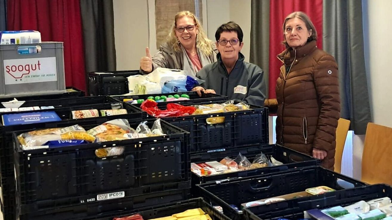 Three women stand next to boxes of food, smiling. One woman gives a thumbs up, another wears glasses. The woman on the right wears a brown coat.