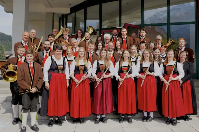 A group of musicians in traditional attire pose for a photo outside a building. Some hold instruments like flutes and tubas, while others wear formal attire. The group includes men and women.