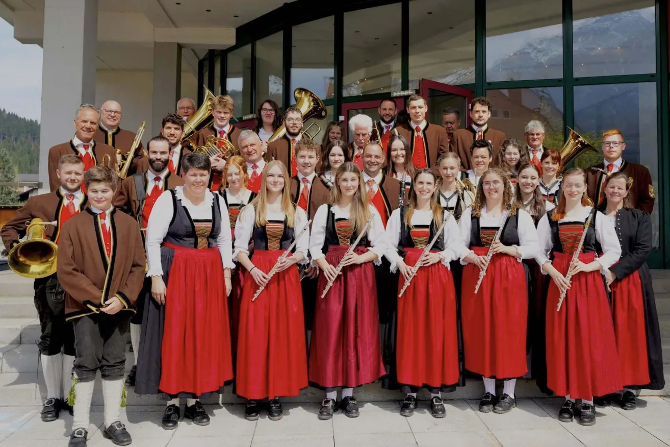 A group of musicians in traditional attire pose for a photo outside a building. Some hold instruments like flutes and tubas, while others wear formal attire. The group includes men and women.