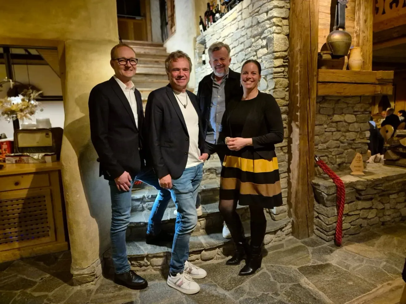 Four people stand in front of a stone wall, smiling. A man on the left wears glasses and a suit, while the man in the middle wears a necklace. The man on the right wears a skirt, and the woman wears a black top.