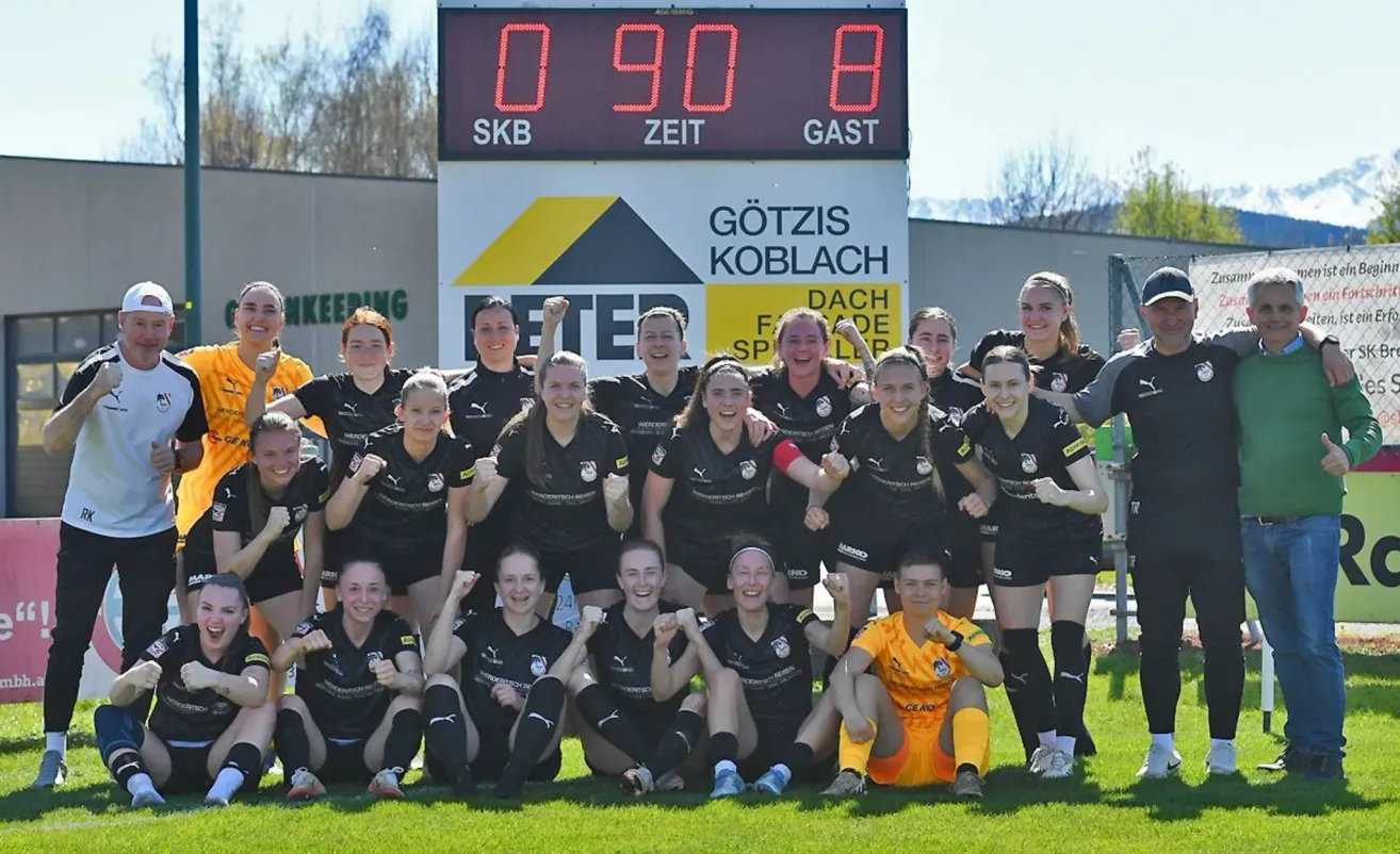 A women's soccer team celebrates in front of a scoreboard that reads 0-90-8, with players in black and yellow jerseys posing for a team photo.