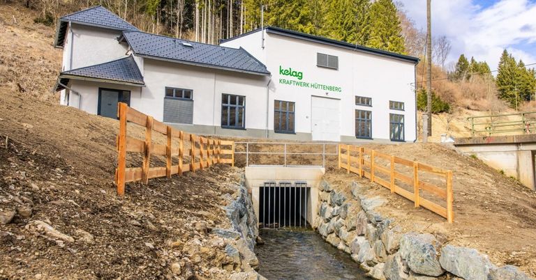 A modern white building with a black roof and the text 'kelag Kraftwerk Huttenberg' is under construction. A wooden fence surrounds the area, and a stream runs through the site.