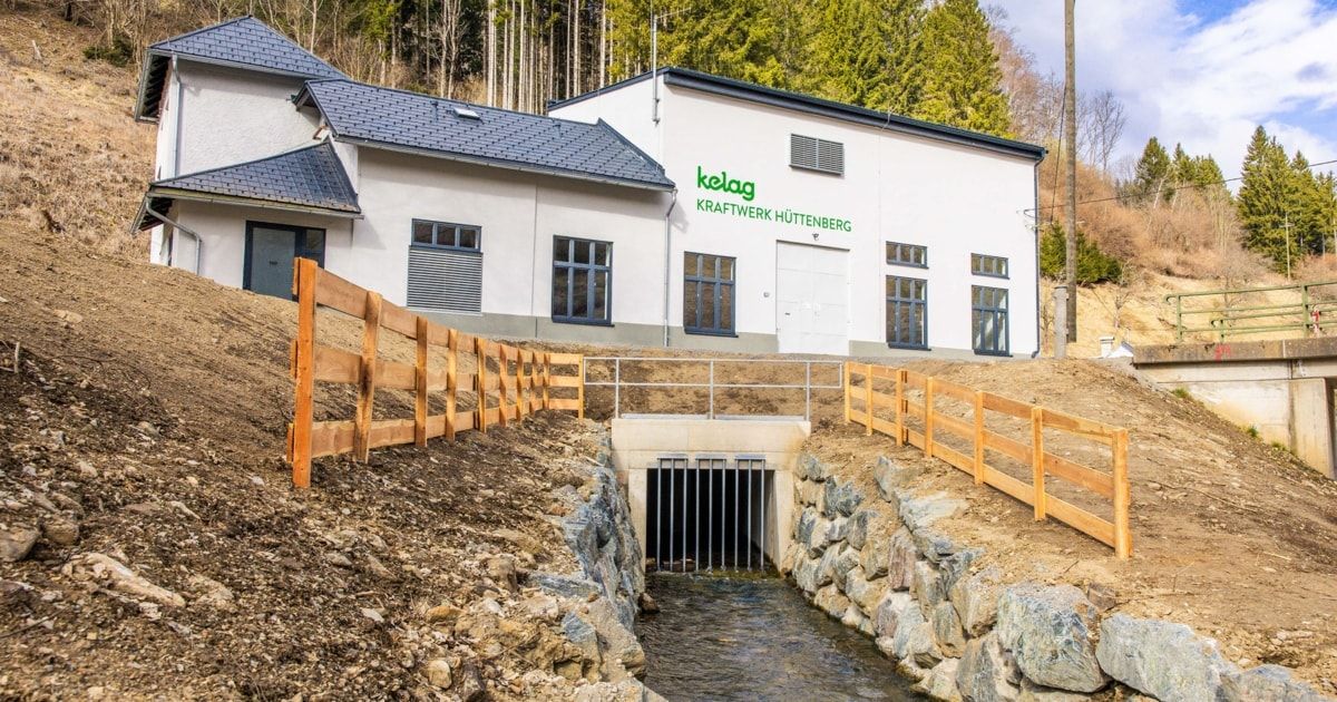 Ein modernes weißes Gebäude mit schwarzem Dach und der Aufschrift 'kelag Kraftwerk Huttenberg' ist im Bau. Ein Holzzaun umgibt das Gelände, und ein Bach fließt durch die Baustelle.