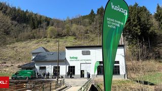 Ein weißes Gebäude mit dem Wort 'kelag' steht vor einem grünen Hügel. Ein grüner Banner steht davor. Menschen stehen vor dem Gebäude.